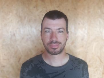 Man with short hair and beard wearing a dark patterned t-shirt against a wooden background.