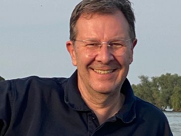 Smiling man wearing glasses and a dark polo shirt outdoors near water.