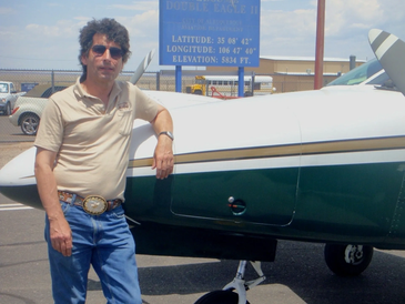 Man in sunglasses leaning on a small airplane with a welcome sign in the background.