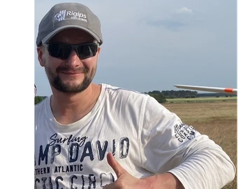 Man wearing sunglasses and a cap giving a thumbs-up outdoors near a glider.