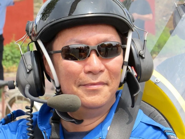 Pilot in blue jumpsuit with helmet and headset smiling.