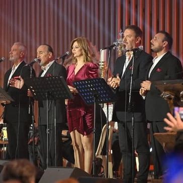 Five people singing on stage with microphones and music stands.
