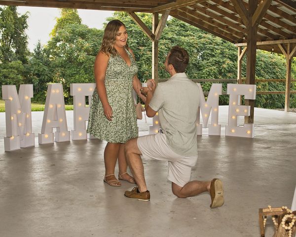 Man proposing to woman under a pavilion with lit MARRY ME letters.