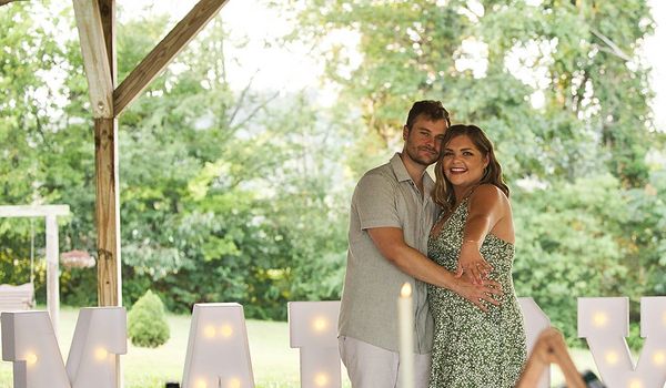 Couple embracing under a pavilion with illuminated 'Marry Me' sign behind them.