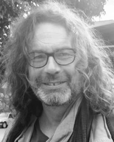 Smiling man with long curly hair and glasses in an outdoor setting.