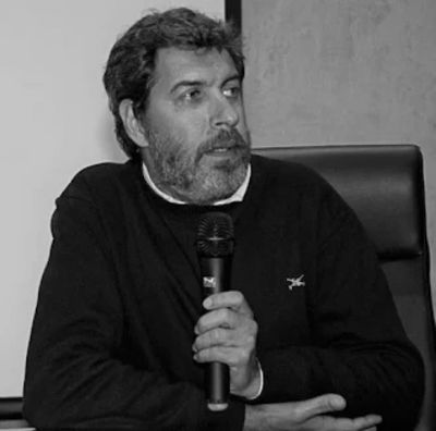 Man with beard holding a microphone, speaking while seated.