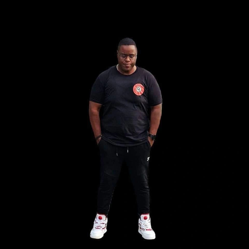 Man in black casual outfit with white sneakers stands against a black background.