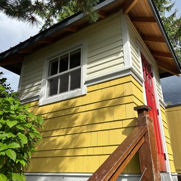 Small yellow and white shed with a bright red door surrounded by greenery.
