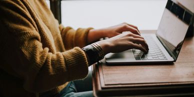 Person typing on a laptop at a wooden desk near a window.