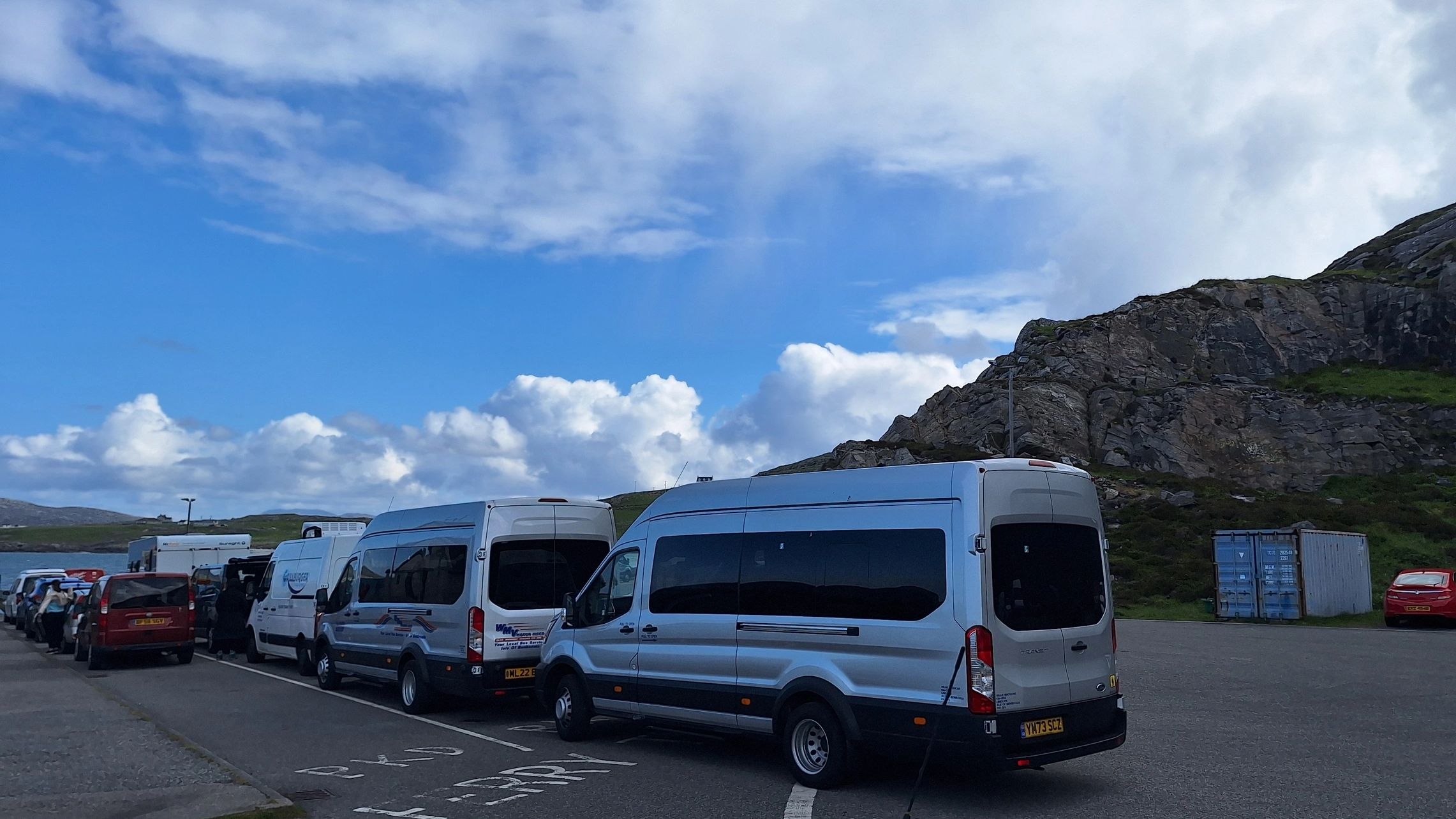 A line of vans and cars parked near rocky hills under a partly cloudy sky.