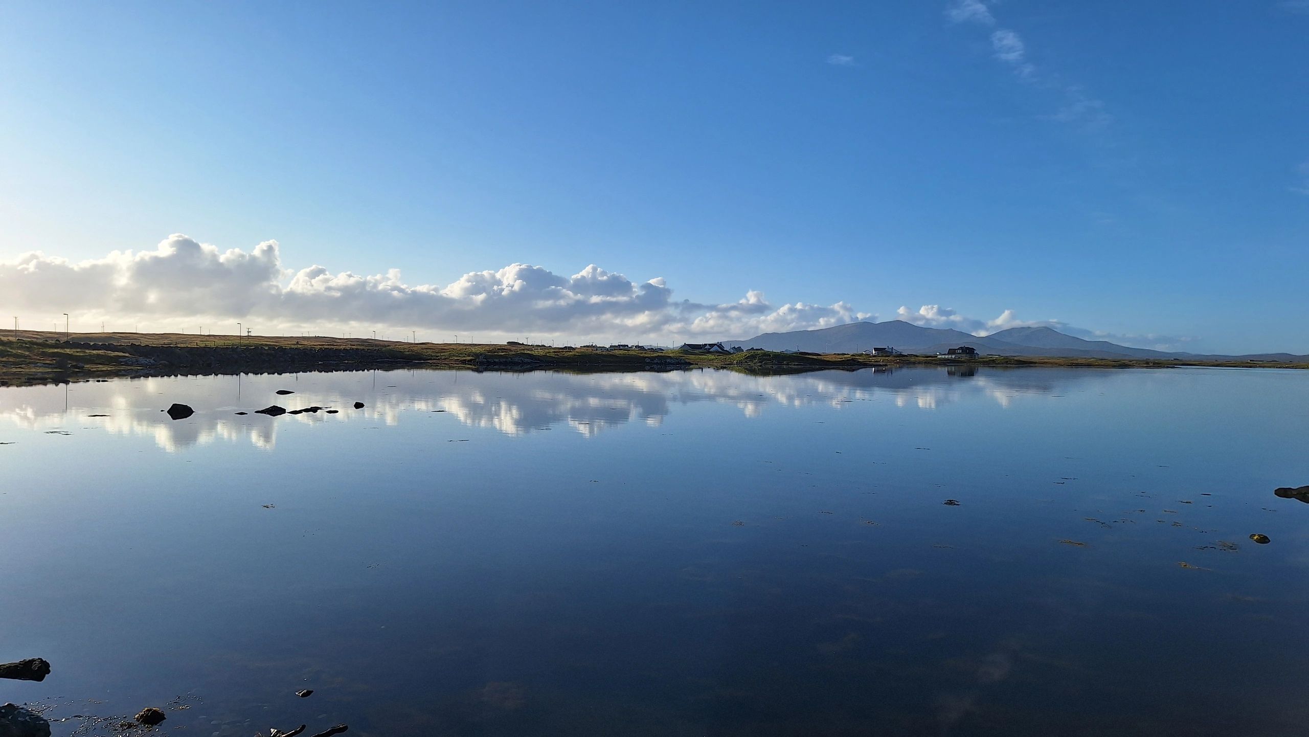 Calm lake reflecting clouds and distant hills under a clear blue sky.