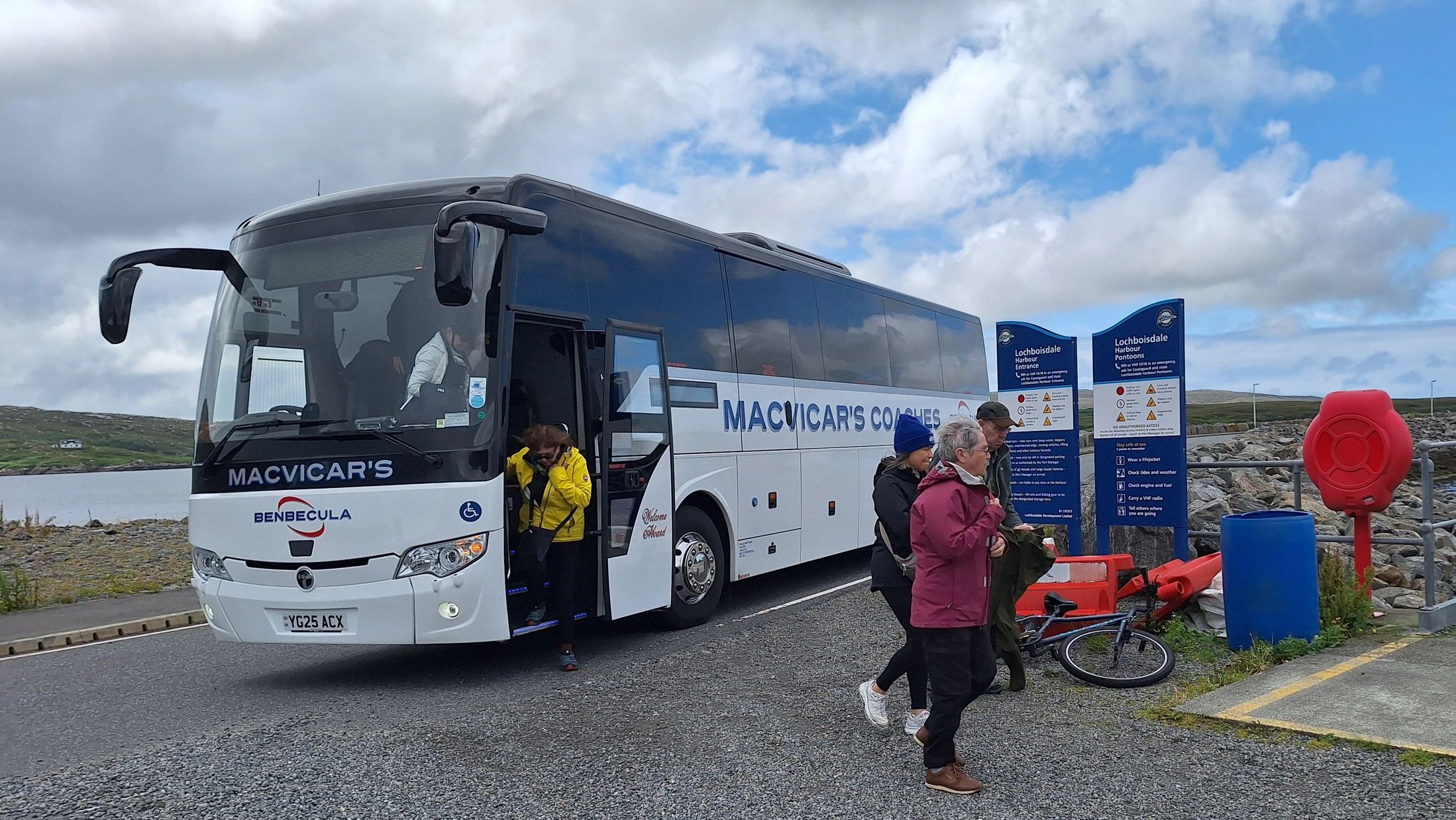 Tour bus at Lochboisdale Harbour with passengers disembarking on a cloudy day.