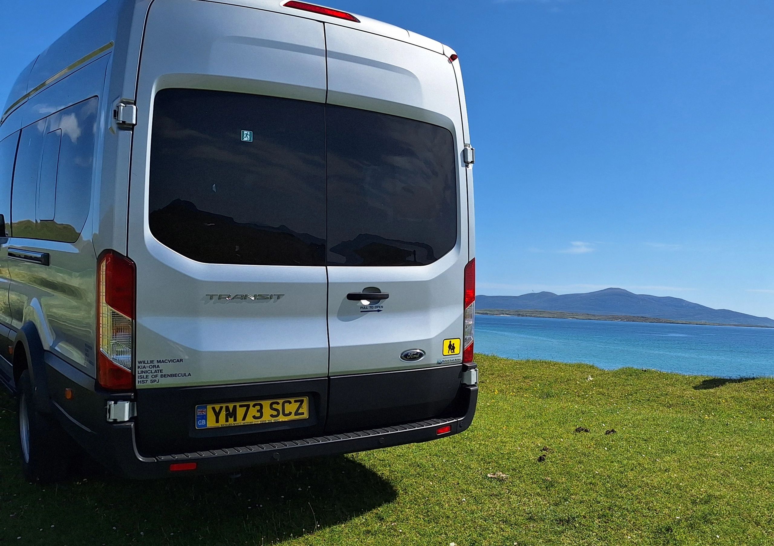 Rear view of a white Ford Transit van parked on grass near a blue sea under clear skies.