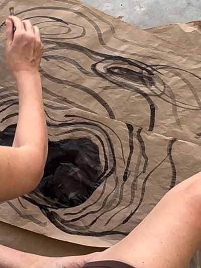 Person painting abstract black swirls on brown paper.
