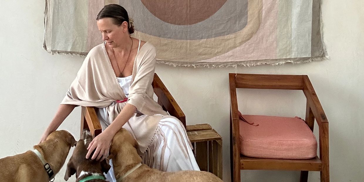 A woman sitting and petting three dogs indoors with a wooden chair beside her.