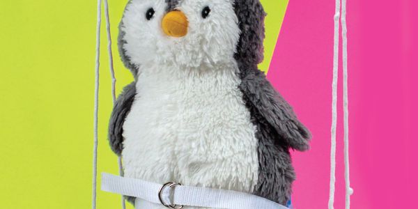 A plush penguin toy on a white swing against a bright yellow and pink background.
