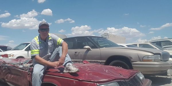 Man sitting on crushed red car in a junkyard.