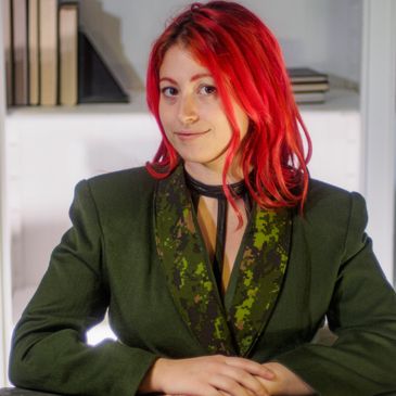 Young woman with vibrant red hair wearing a green blazer, sitting at a desk.