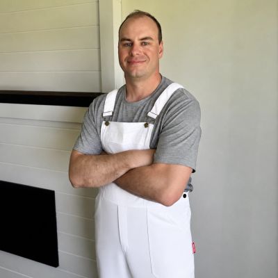 Man in white overalls and gray shirt standing with crossed arms indoors.