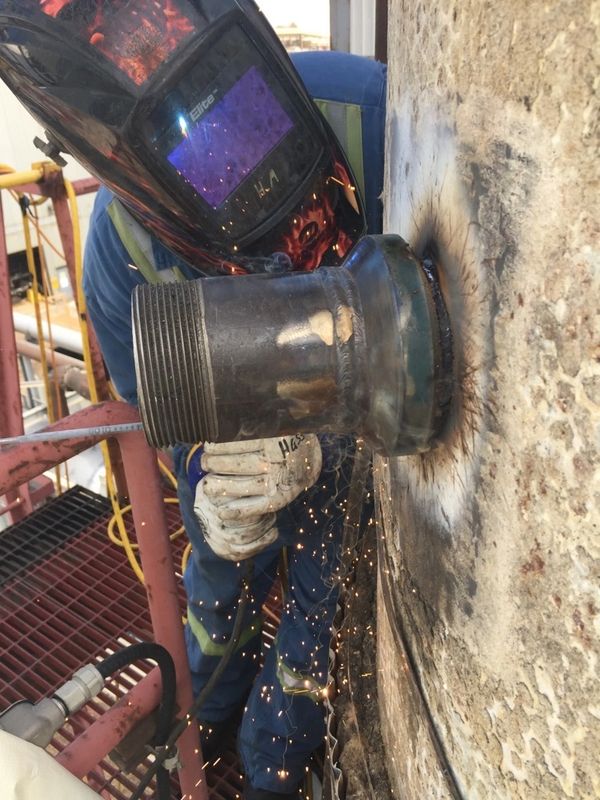 Welder in protective gear welding a pipe to a metal surface with sparks flying.