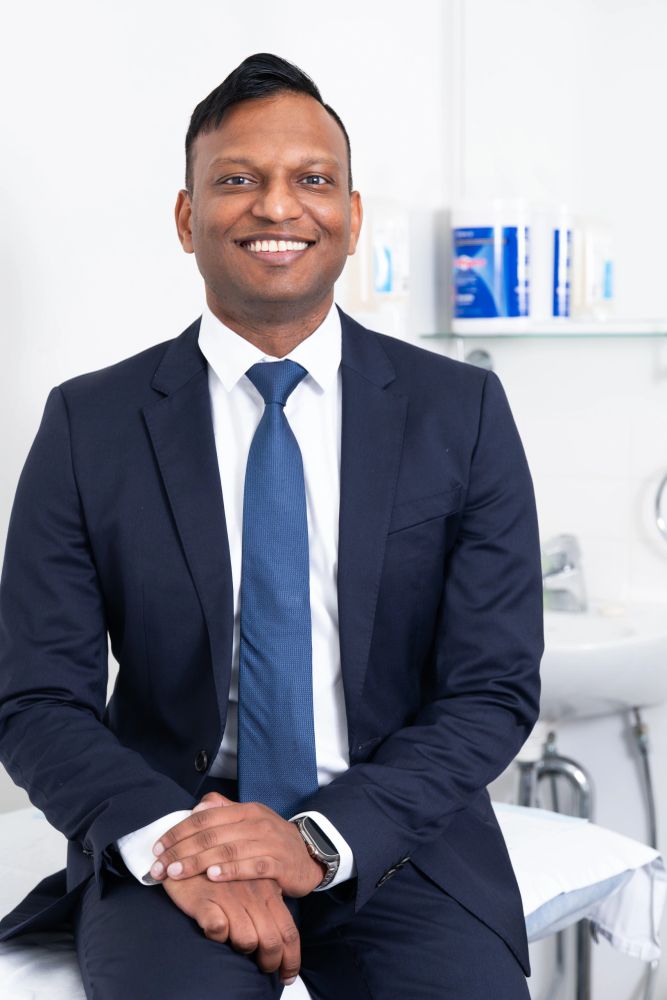 Man in a navy suit smiling warmly, sitting in a medical or clinical setting.