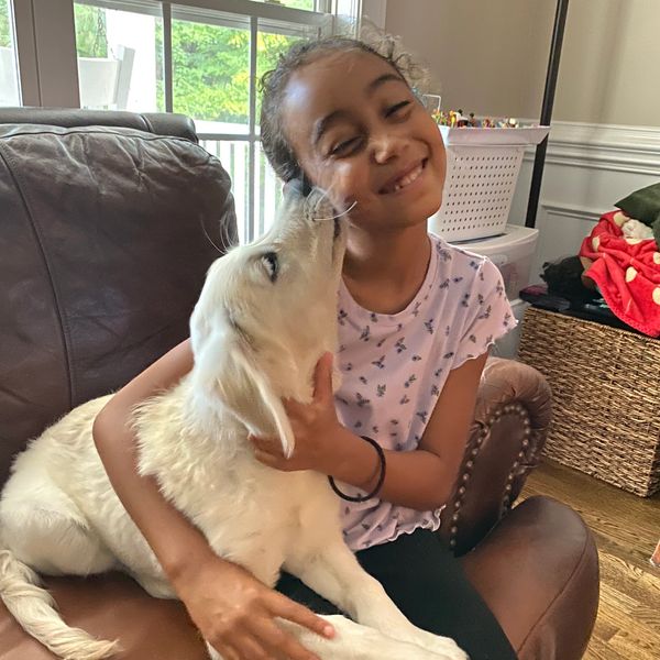A girl happily hugging her dog on a couch as the dog licks her face.
