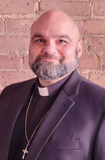 Smiling priest wearing a clerical collar and cross necklace.