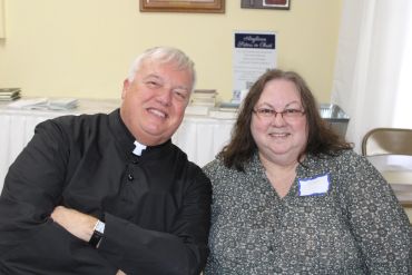 Man in priest attire and woman smiling indoors.