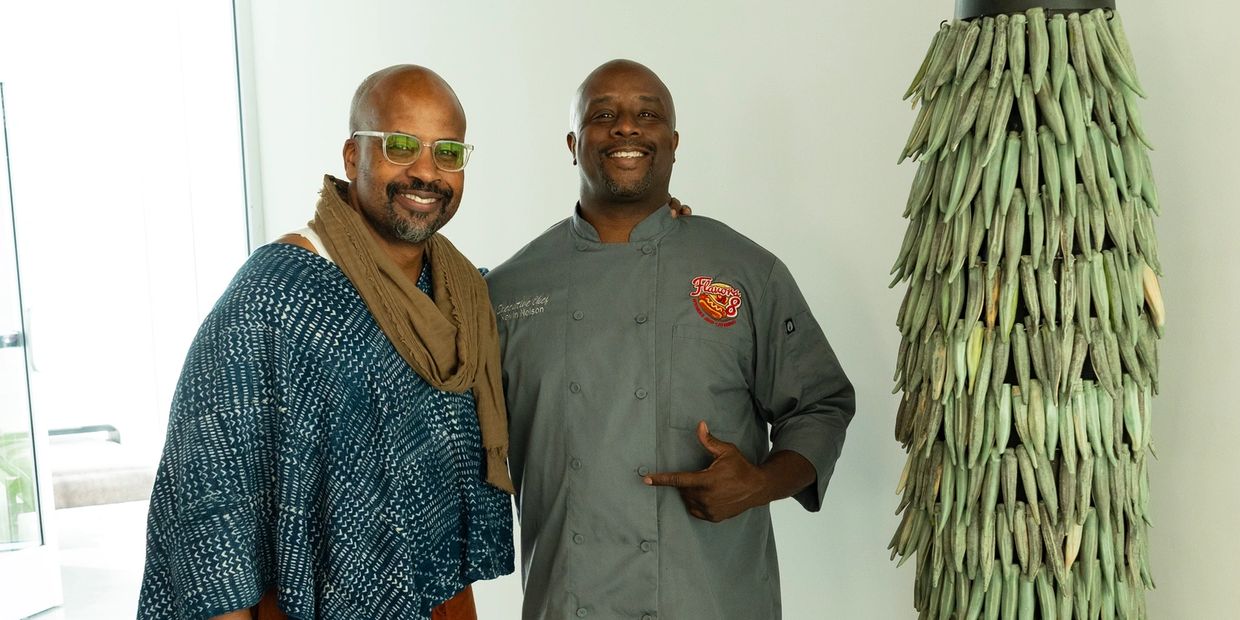 Two men smiling, one wearing chef outfit, next to a tall art piece.