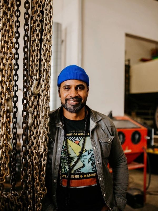 Man wearing a blue beanie and graphic t-shirt stands next to hanging chains in a workshop.