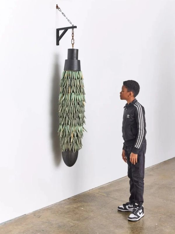 Boy observing a hanging sculpture with green leaf-like elements in a gallery.