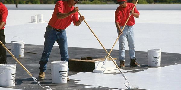 Workers in red shirts applying white coating on a flat roof with rollers.