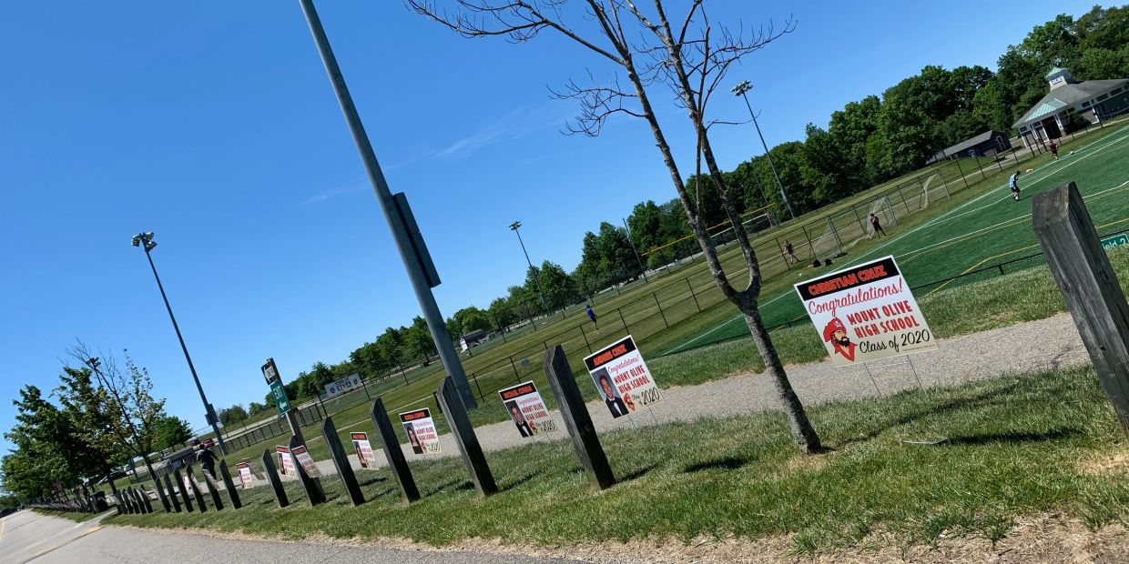 Yard signs congratulating Mount Olive High School Class of 2020 line a pathway beside a sports field.