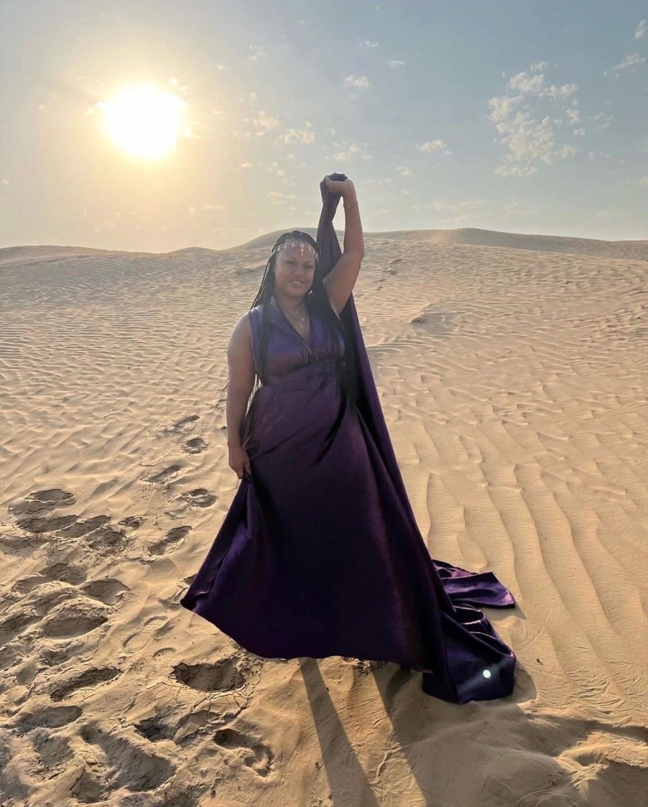 Woman in a flowing purple dress poses on sunlit sand dunes.