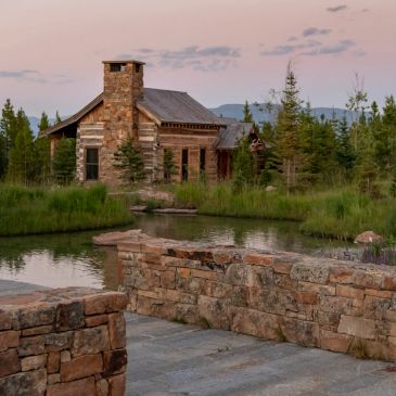Rustic cabin next to a pond with stone walls in the foreground.