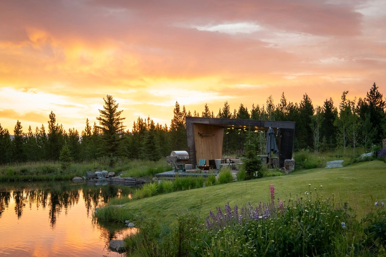 Sunset over a lakeside patio with vibrant flowers and pine trees.