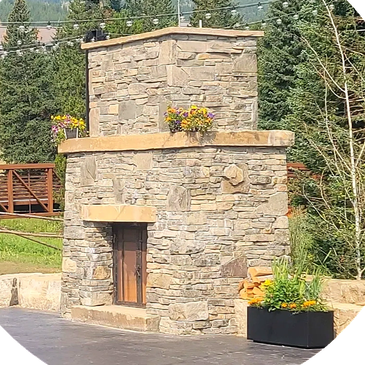 Stone fireplace outdoors with flowers on top and nearby.