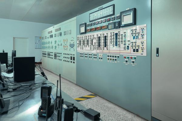 Control room with large electrical panels, monitors, and communication equipment on a desk.