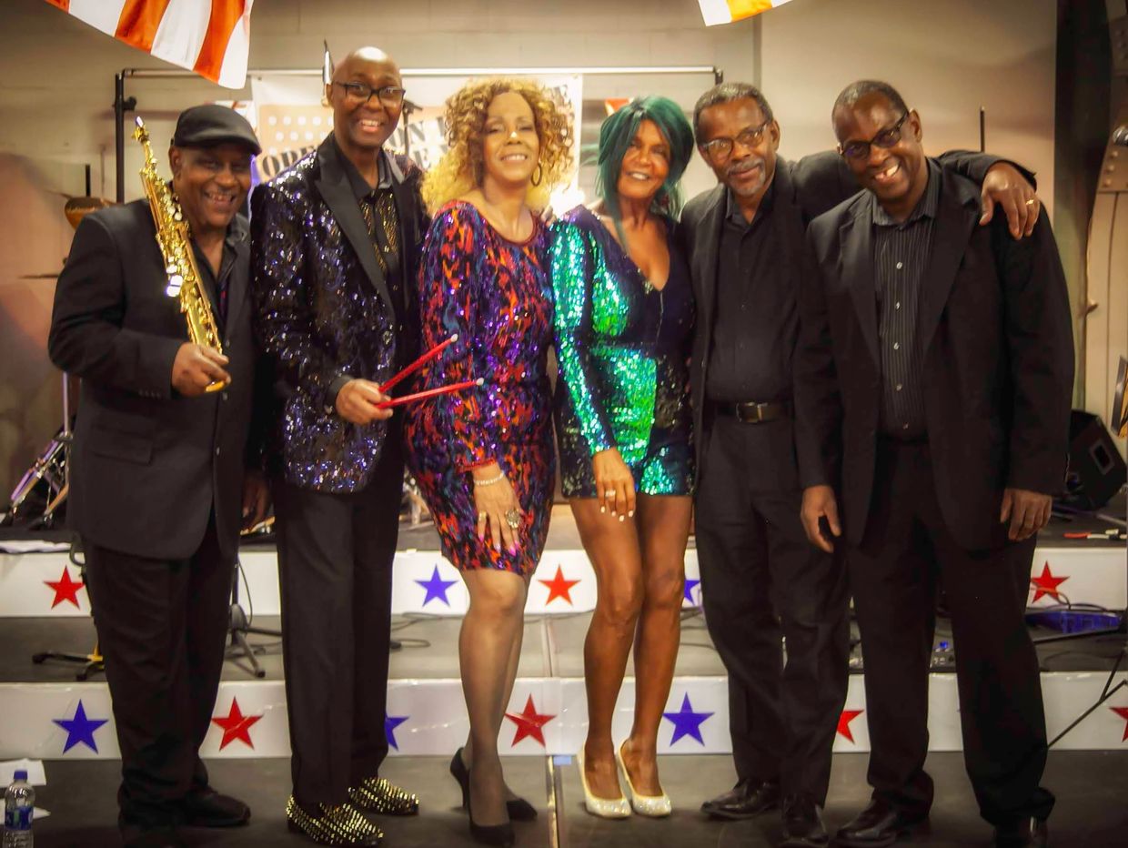 A group of six musicians posing together on stage in colorful and black attire.