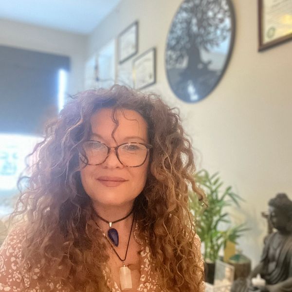 Woman with curly hair and glasses smiling indoors with plants and artwork in the background.