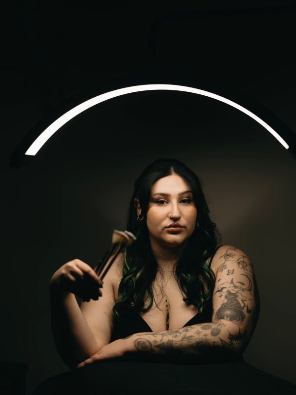 Woman with tattoos holding makeup brushes under a glowing semicircle light.