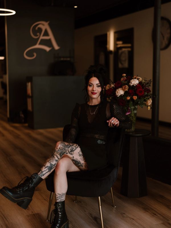 Tattooed woman in black outfit sitting confidently in a modern room with flowers.