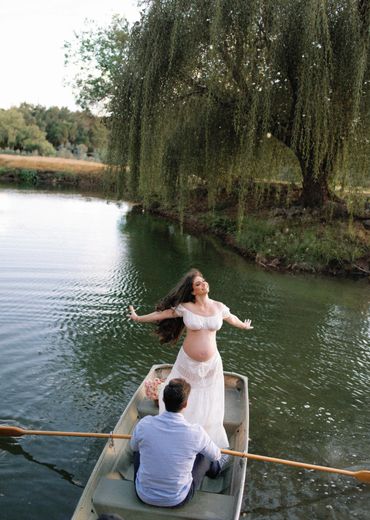 A pregnant woman stands gracefully at the front of a boat on a peaceful lake.