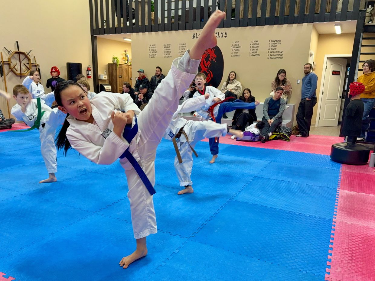 Young martial artists practicing high kicks in a dojo with spectators watching.
