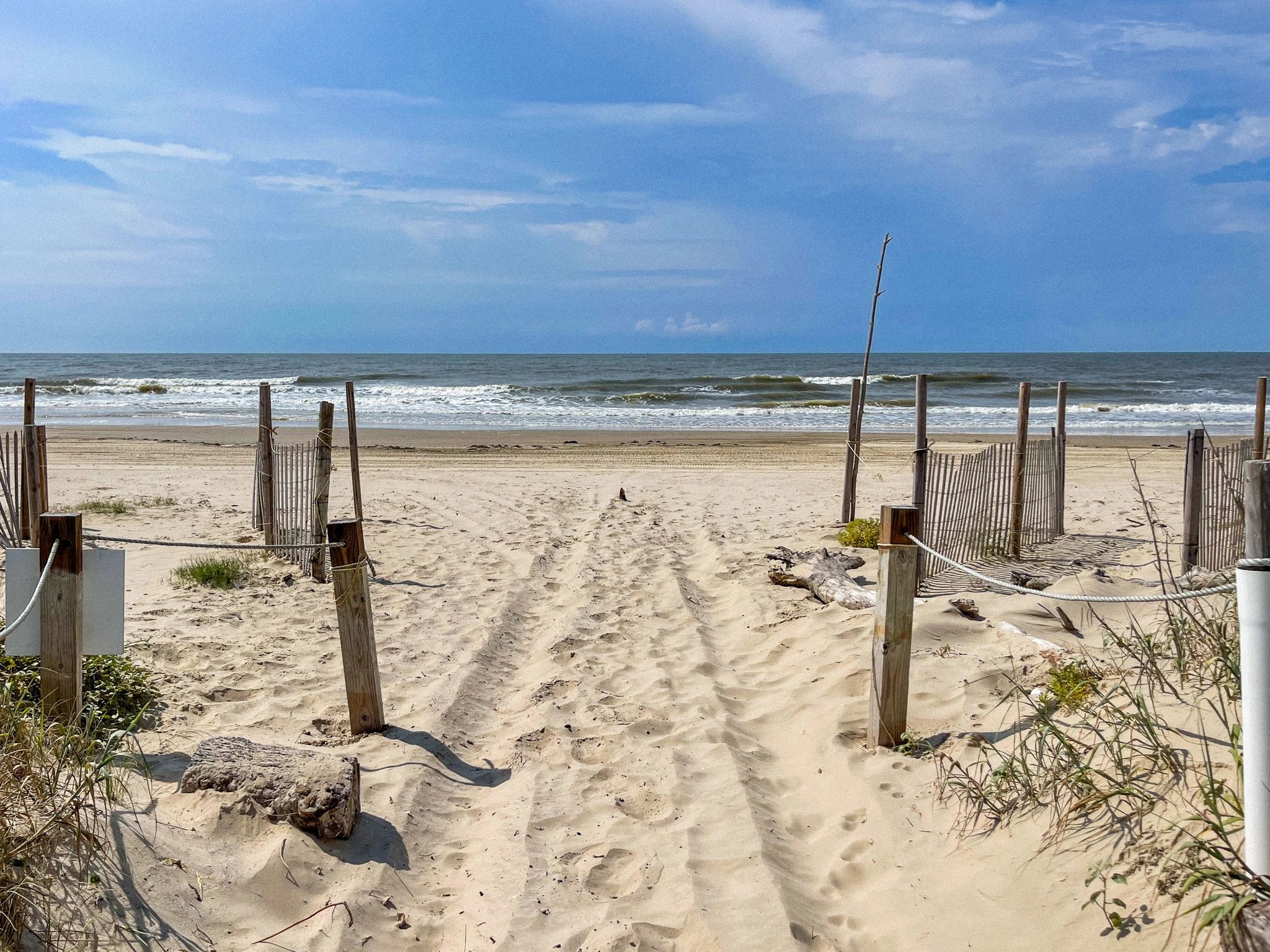Sandy beach pathway leading to the ocean with wooden fences and blue sky.