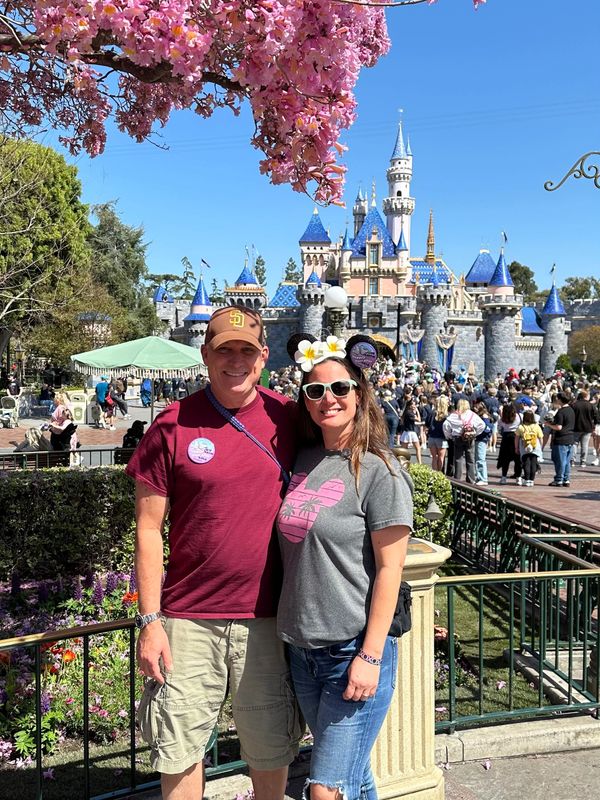 Couple posing happily in front of Disneyland's Sleeping Beauty Castle with cherry blossoms above.