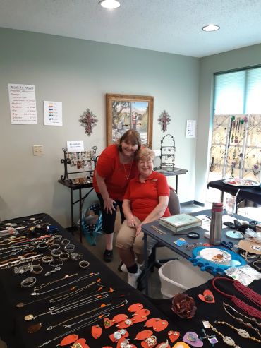 Two women smiling at a jewelry sale event with tables full of necklaces and bracelets.