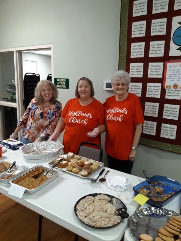Three women smiling behind a table of baked goods at a church event.