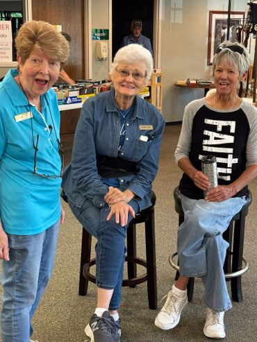 Three elderly women smiling and posing indoors, one wearing a faith shirt.