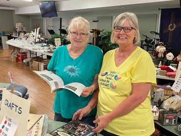 Two smiling elderly women at a crafts sale table indoors.